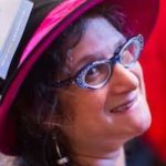 Smiling, middle aged white woman in a black hat with a dark pink brim, wearing blue cat eye glasses