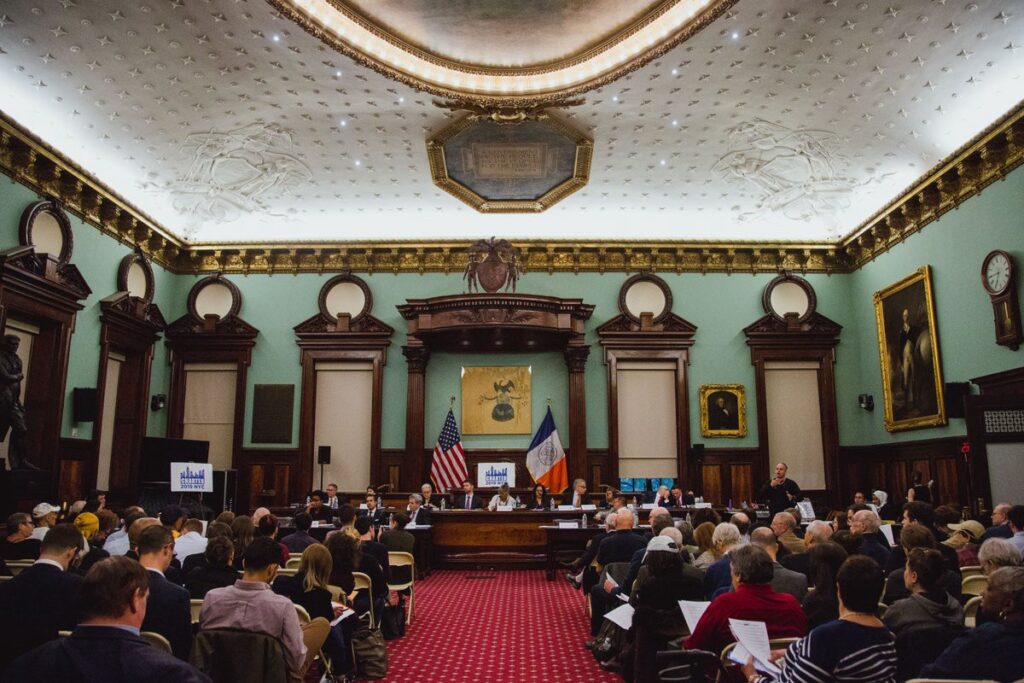 NYC Council chamber, ornate, white ceiling, light blue walls with dark wood window frames and red carpet