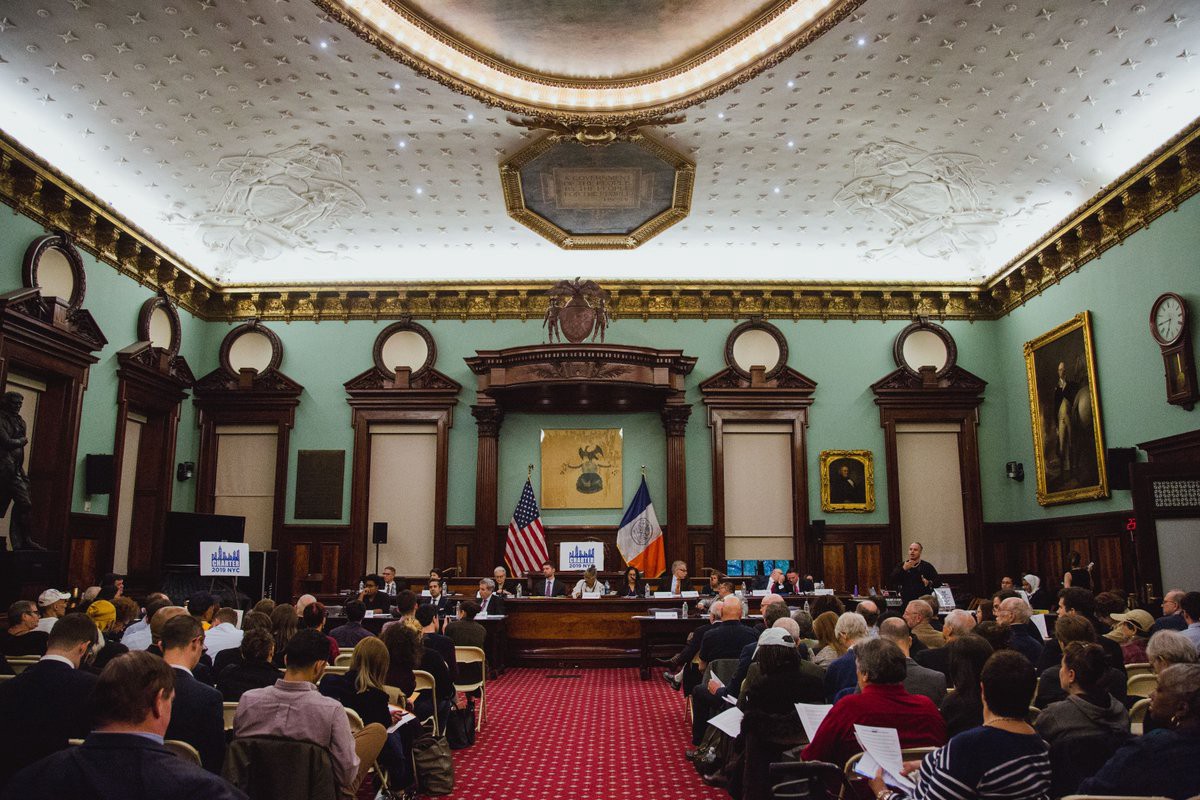 NYC Council chamber, ornate, white ceiling, light blue walls with dark wood window frames and red carpet