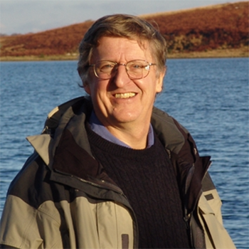 Photo of Michael Goodchild, smiling middle-aged white man wearing glasses, a black sweater and an open khaki jacket. Background is open water with a hill in the distance.